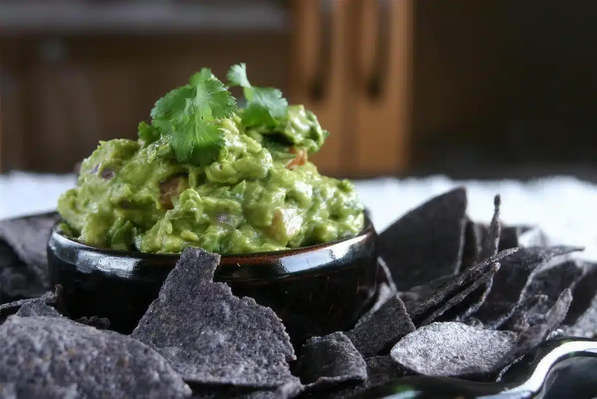 A small dark bowl of avocado dip with chips scattered around it