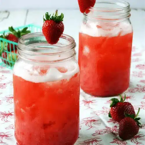 Two tall jars of a pink iced drink beside fresh fruit on a printed cloth