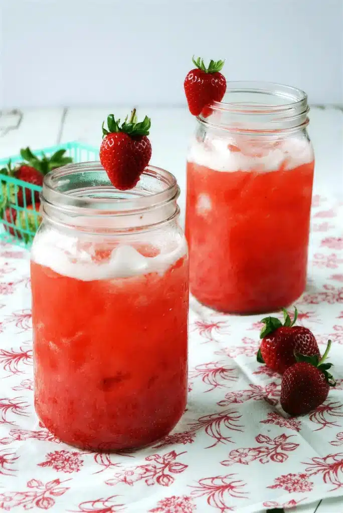 Two tall jars of a pink iced drink beside fresh fruit on a printed cloth
