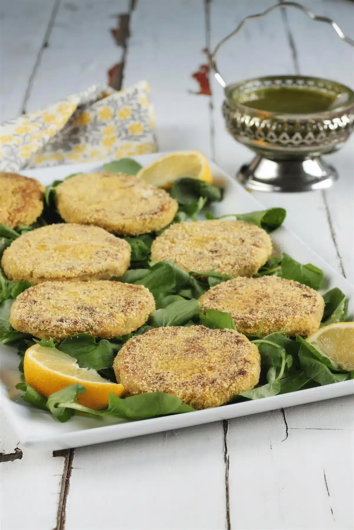 Chickpea patties on a white platter with greens, lemon and a bowl of herb sauce
