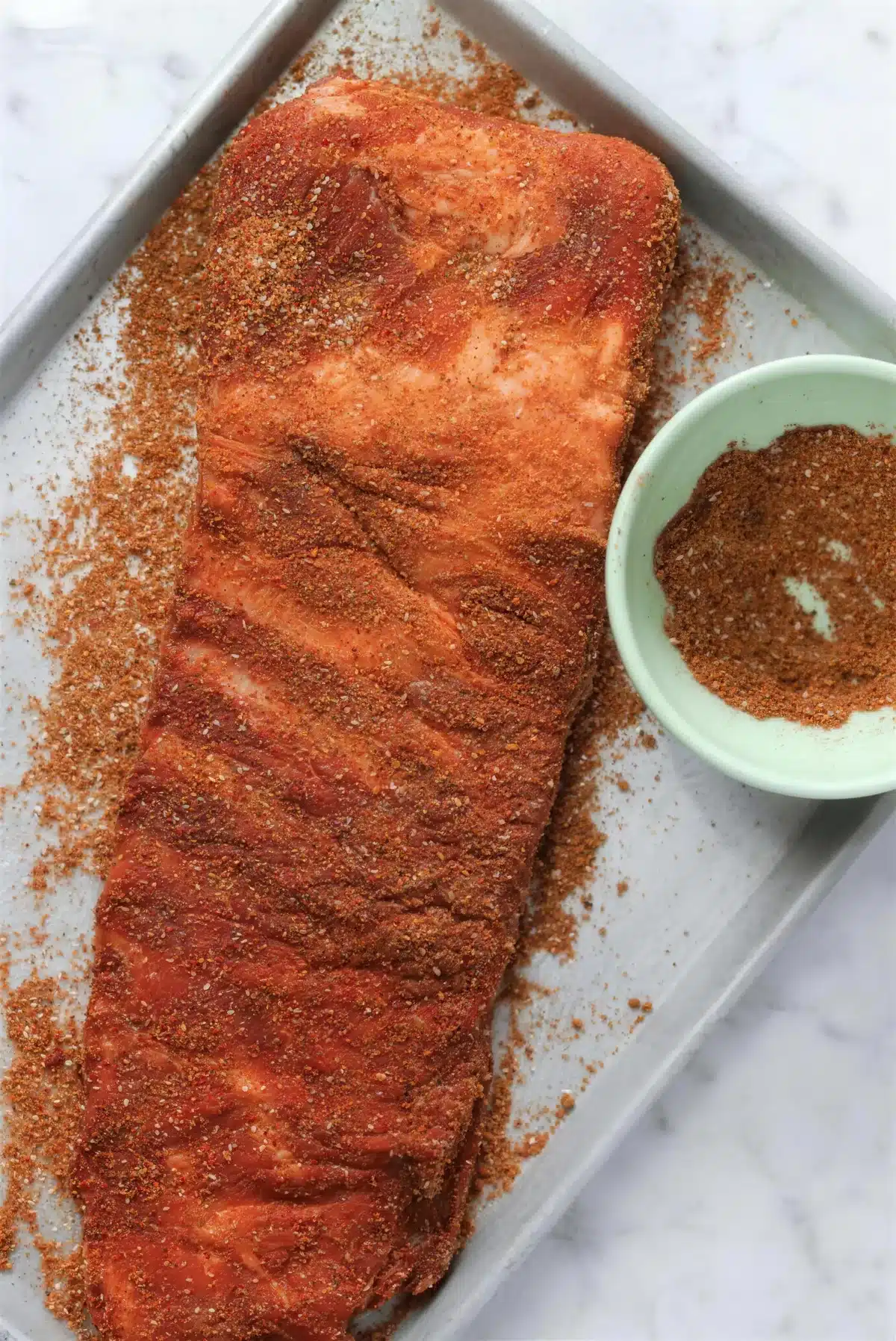 A seasoned rack of pork resting on a sheet pan beside a bowl of ground spice rub