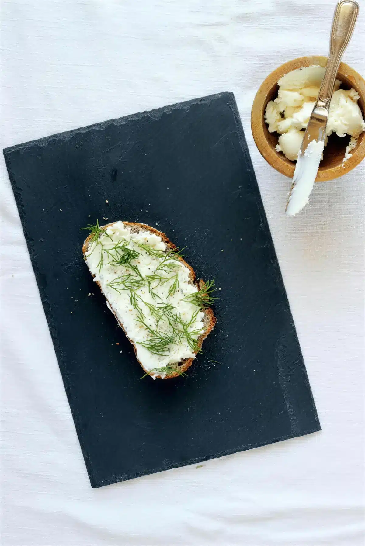 Toasted rustic bread spread with soft white cheese and torn dill on slate