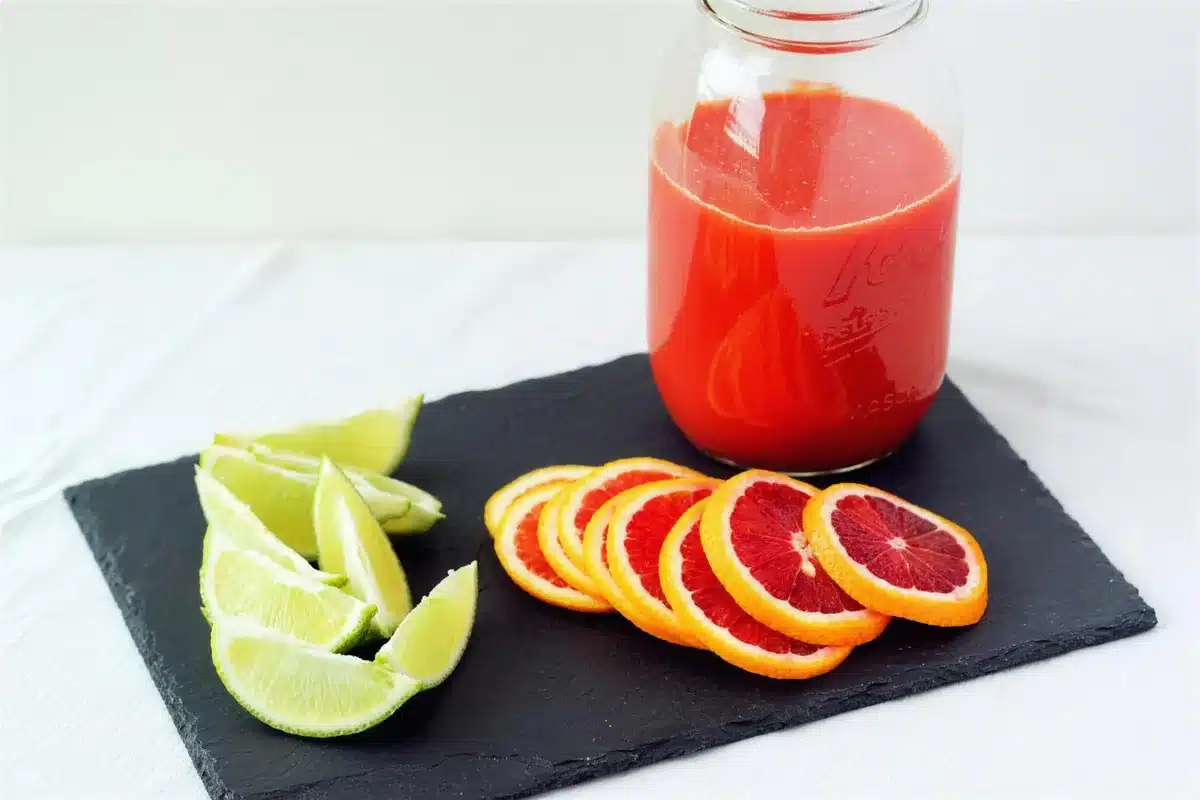 A jug of ruby juice with lime wedges and sliced citrus on slate