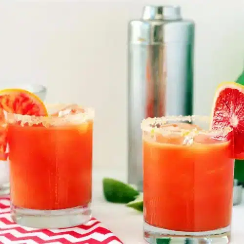 Blood orange cocktail in two glasses beside a shaker and a bottle of ginger beer