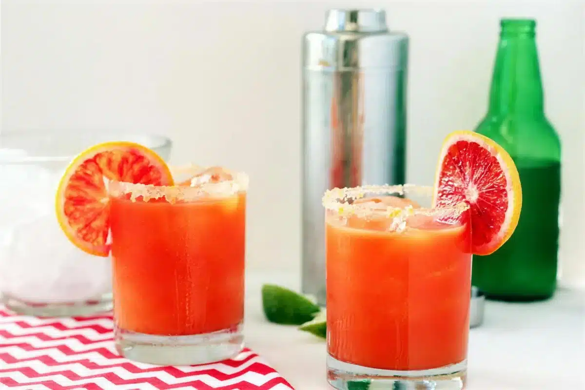 Blood orange cocktail in two glasses beside a shaker and a bottle of ginger beer
