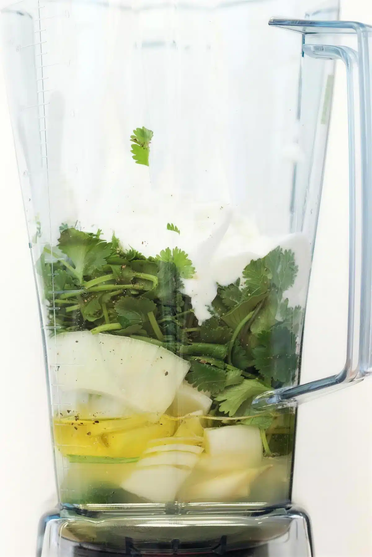 A tall blender jar packed with fresh herbs, onion and yogurt before blending