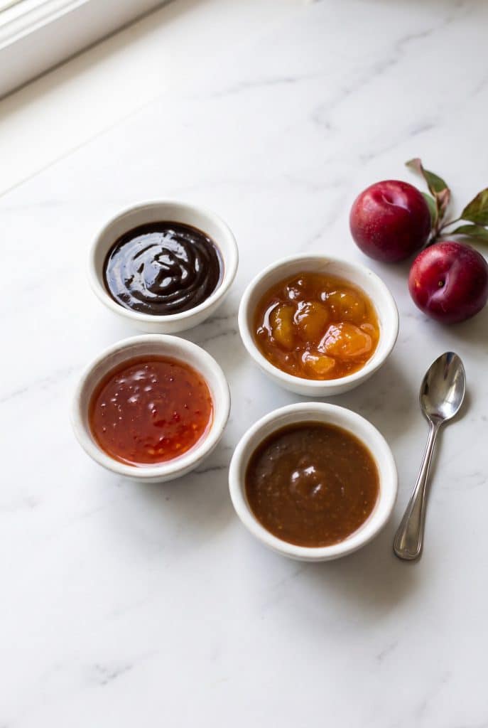 Four small bowls of dark and amber sauces beside fresh red plums