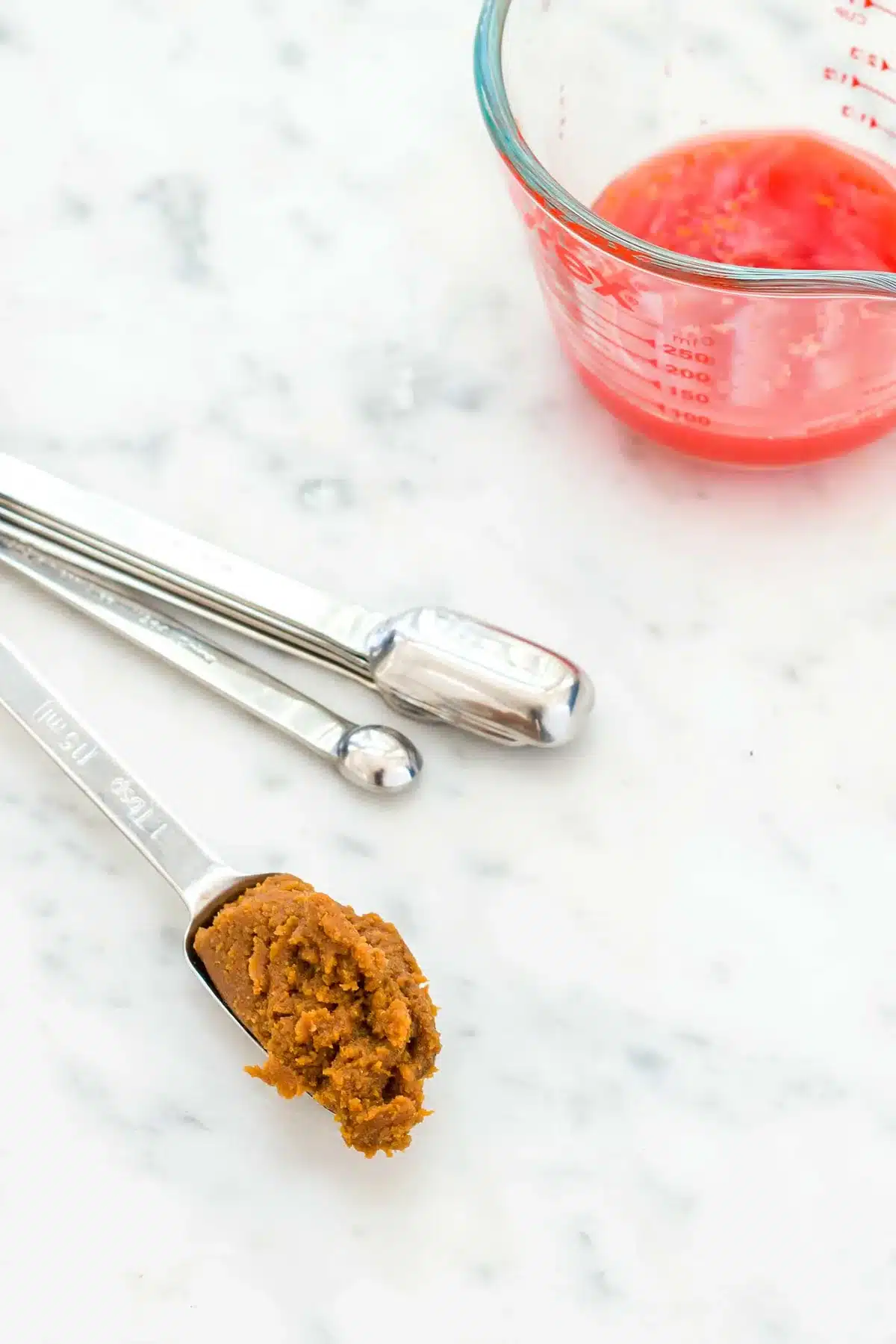 A measuring spoon of dark fermented paste beside a jug of red citrus juice