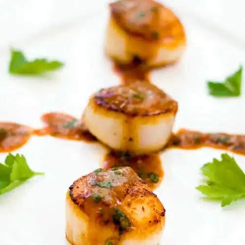 Three golden crusted shellfish rounds stacked on a white plate with glossy rust sauce