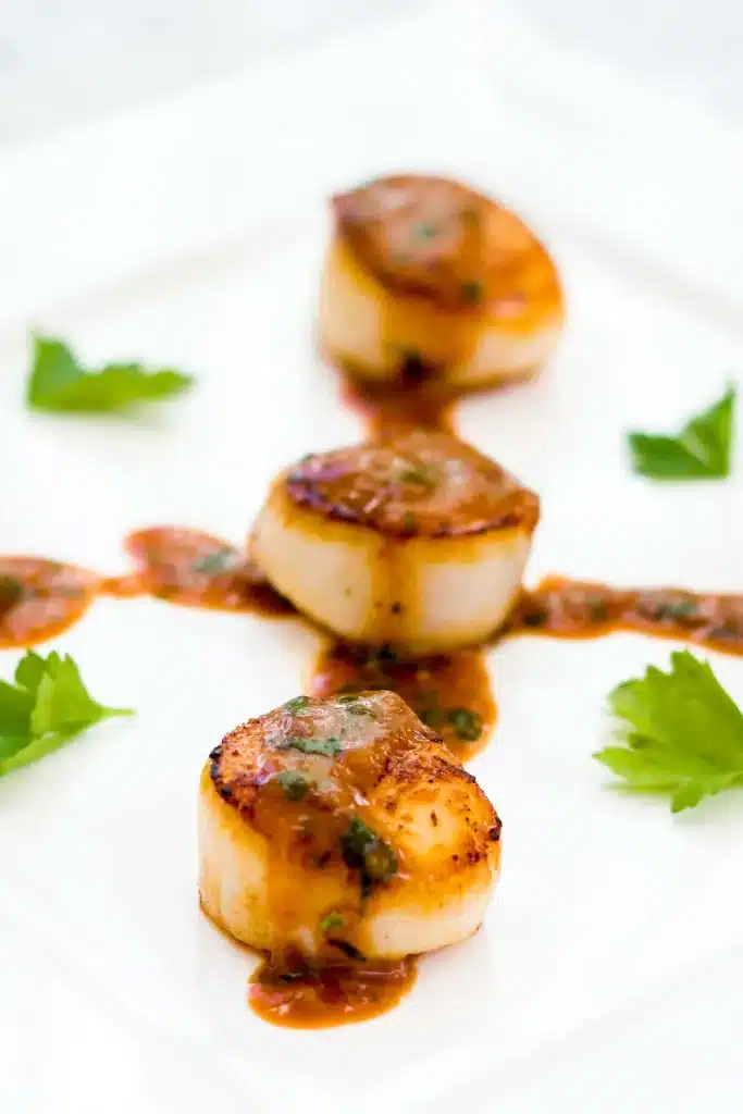 Three golden crusted shellfish rounds stacked on a white plate with glossy rust sauce