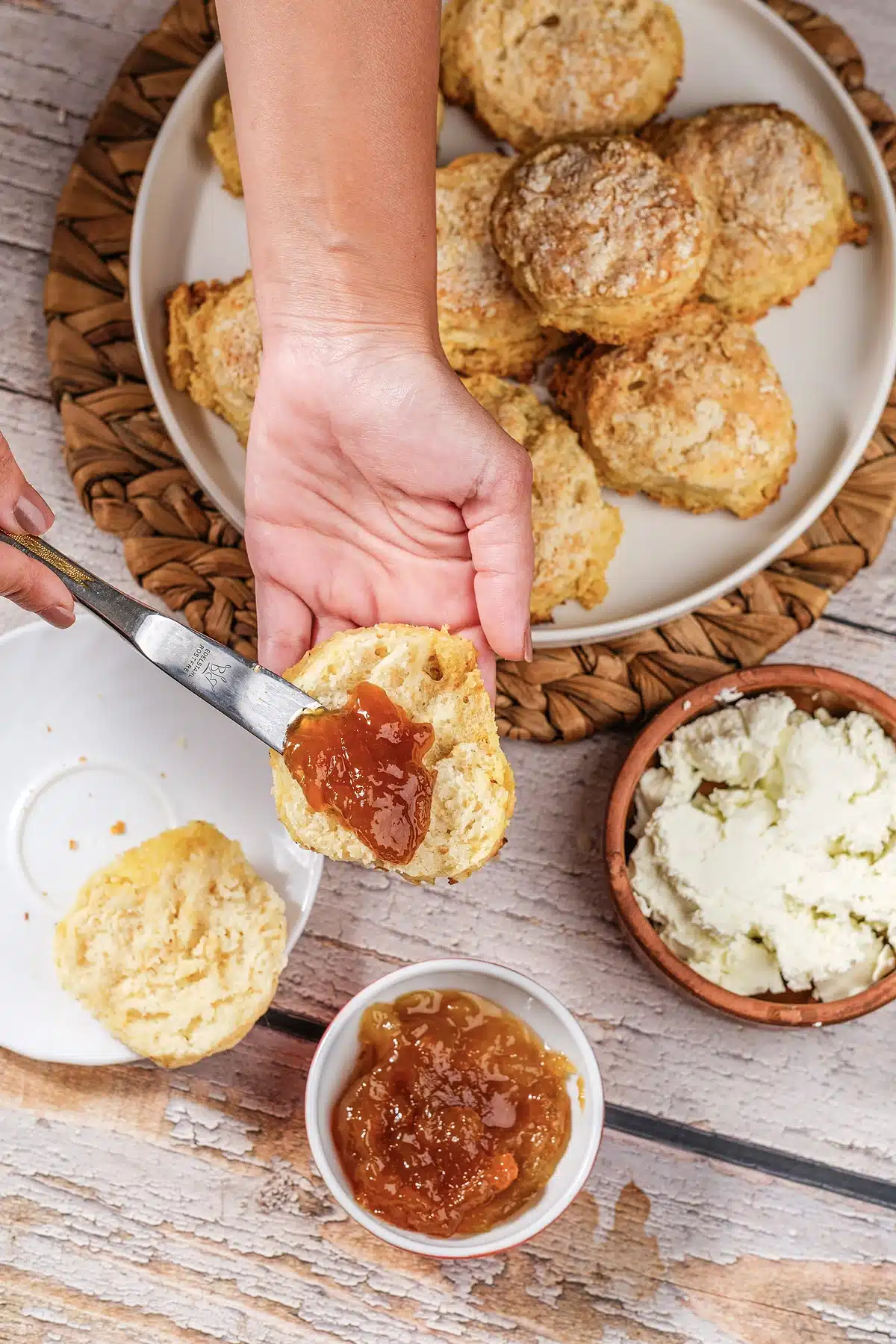A hand spreading jam over a split 7Up biscuit with a bowl of preserves below