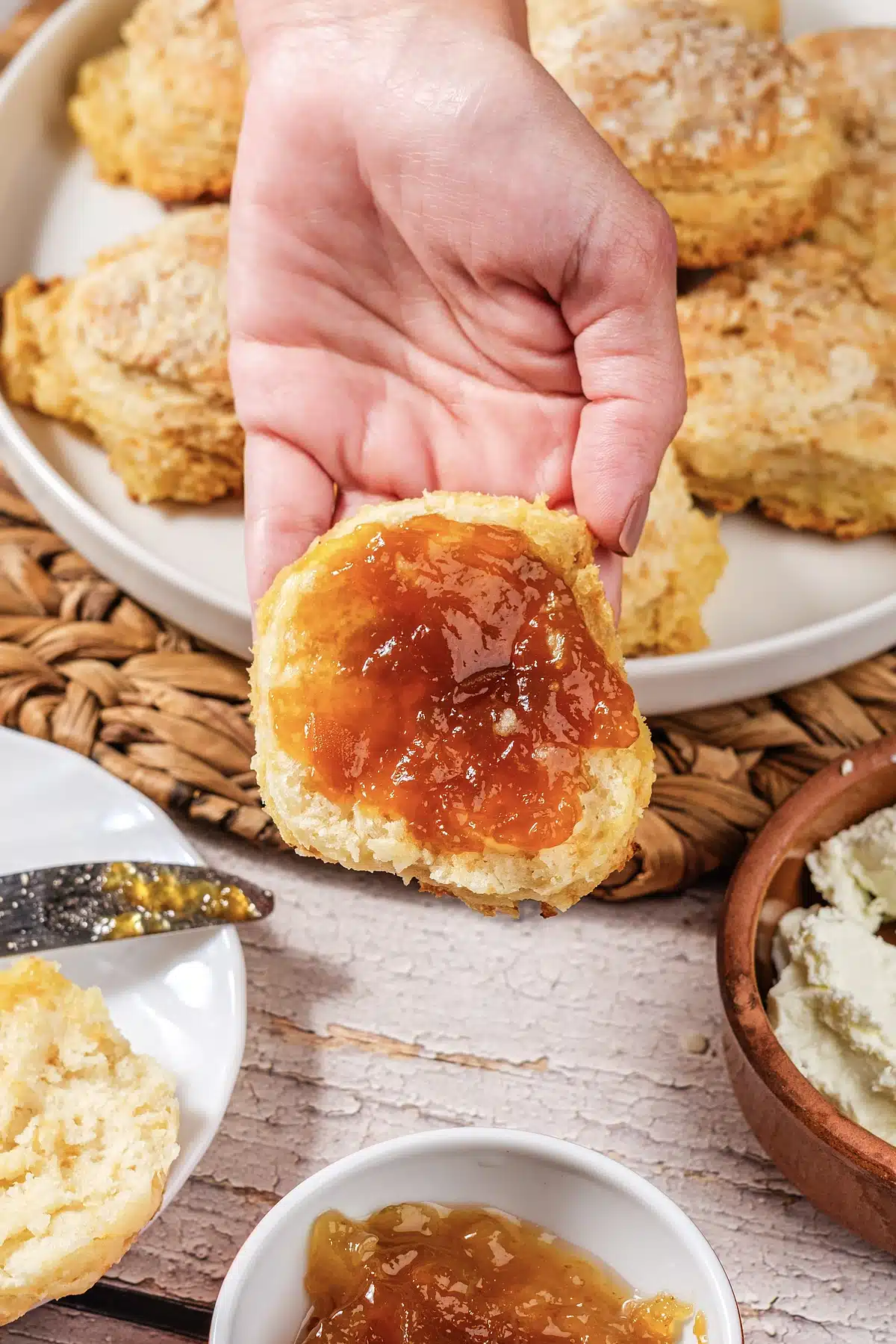 A hand holding a jam-topped round above a plate and a bowl of preserves
