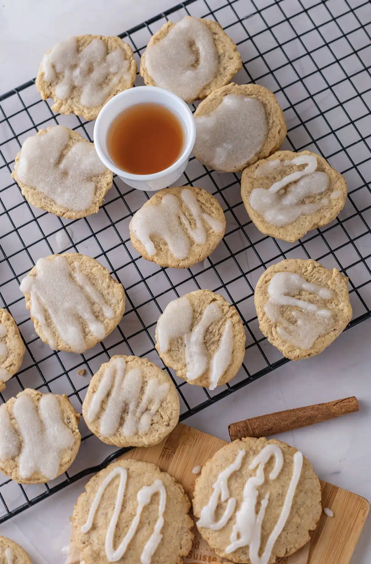 Apple cider cookies on a wire rack and a wooden board, some with a thin glaze and some with a piped zigzag, beside a cup of cider