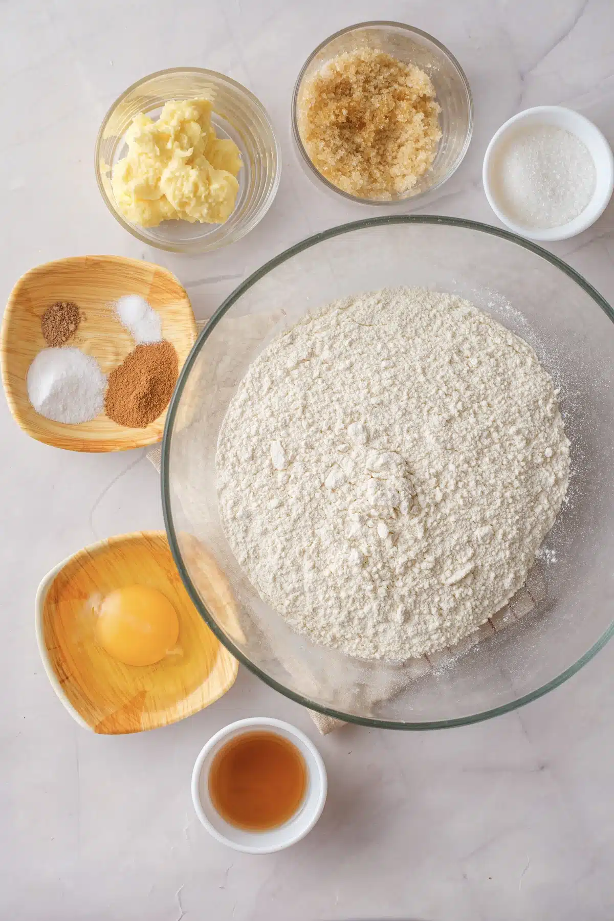 A bowl of flour with butter, brown sugar, granulated sugar, an egg, cinnamon, nutmeg, salt and a small cup of apple cider around it