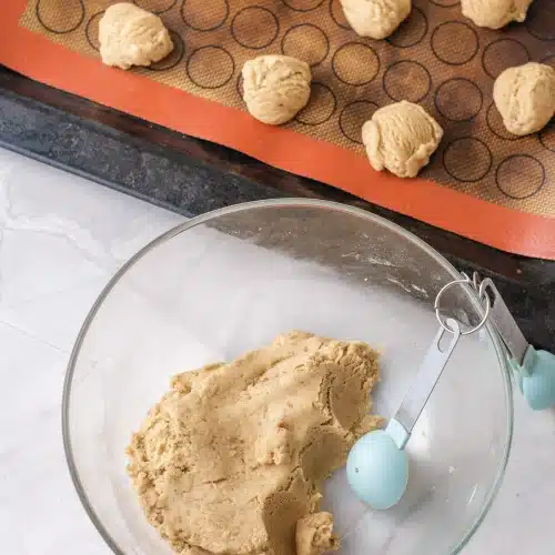 Dough balls scooped onto a silicone-lined baking sheet beside the bowl of dough and a scoop