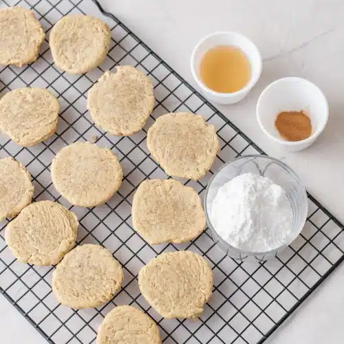 Baked rounds cooling on a wire rack beside small bowls of cider, cinnamon and powdered sugar
