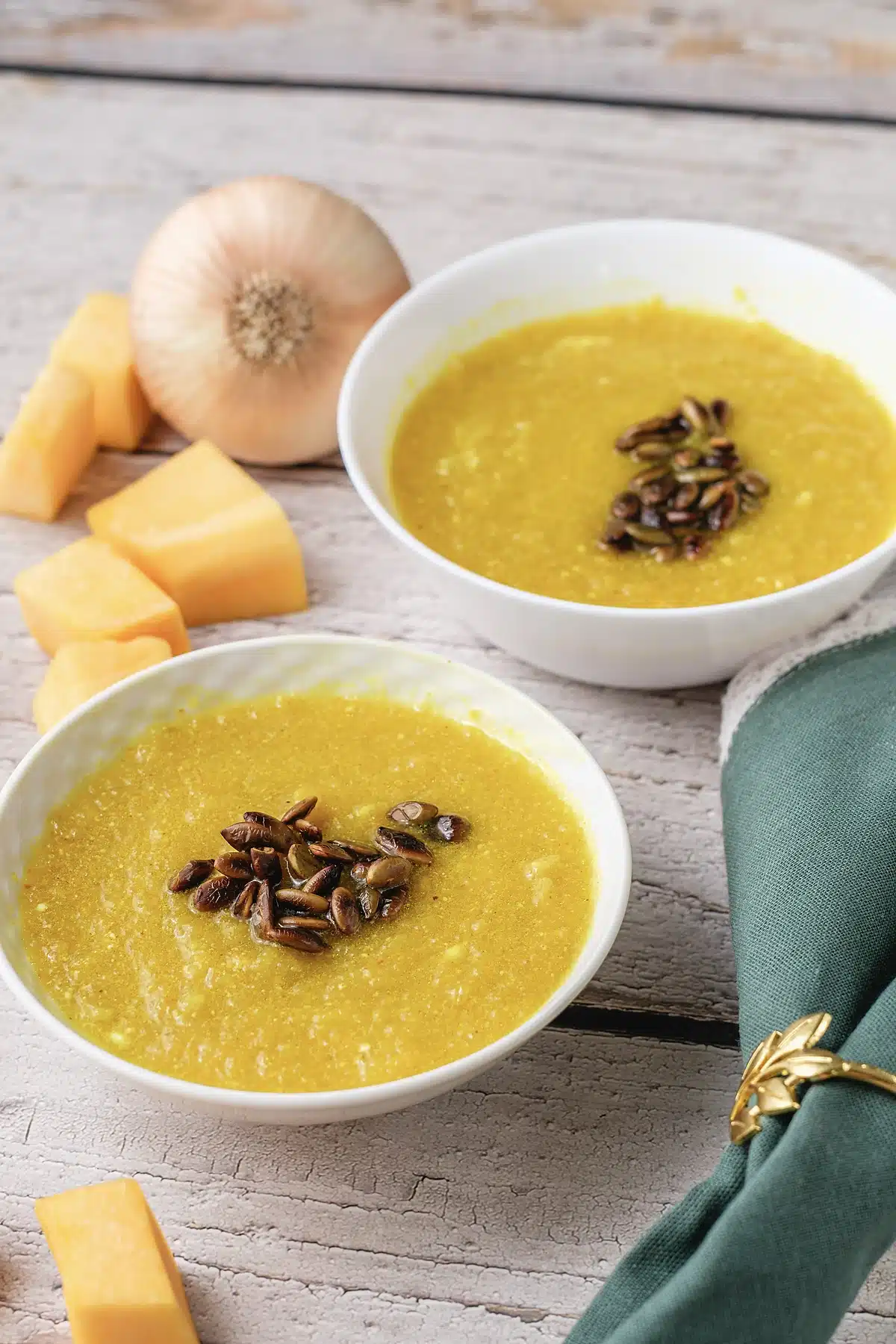 A close bowl of golden soup with pepitas, squash cubes and an onion behind