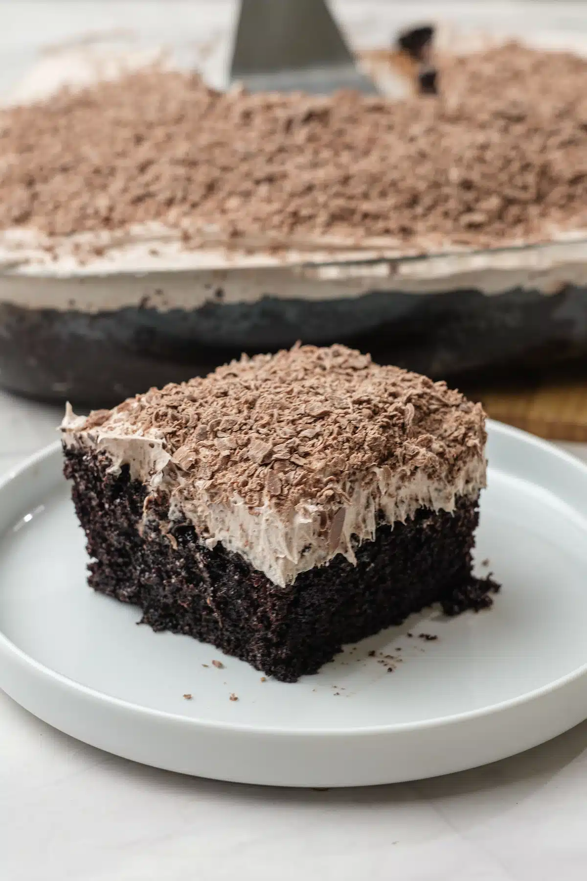 A slice of Baileys poke cake on a white plate showing the dark crumb and thick topping, with the baking dish and a spatula behind