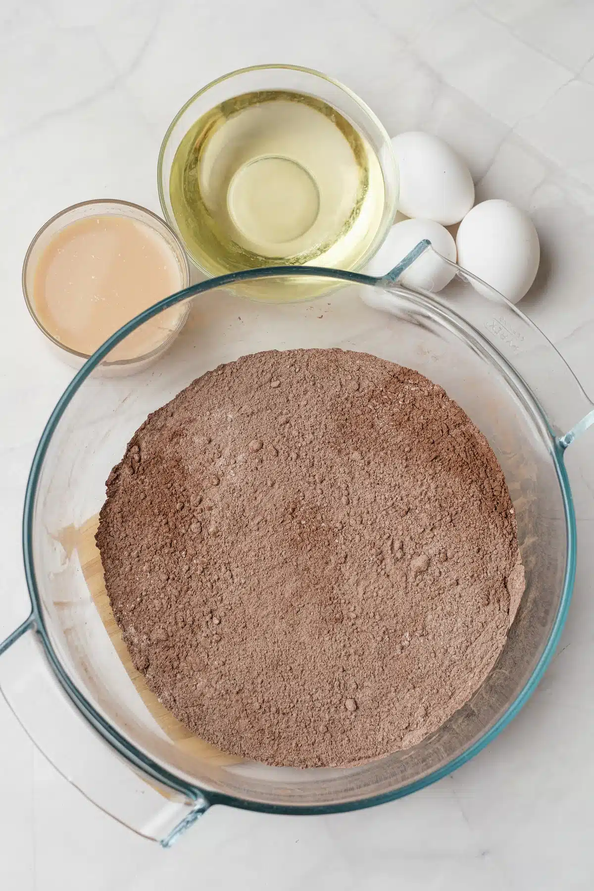 A bowl of chocolate cake mix with oil, eggs and a glass of Baileys beside it on a marble counter