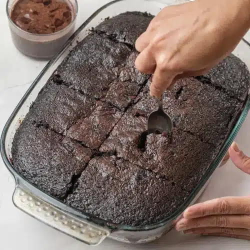 A hand poking holes across the warm cake with the handle of a spoon