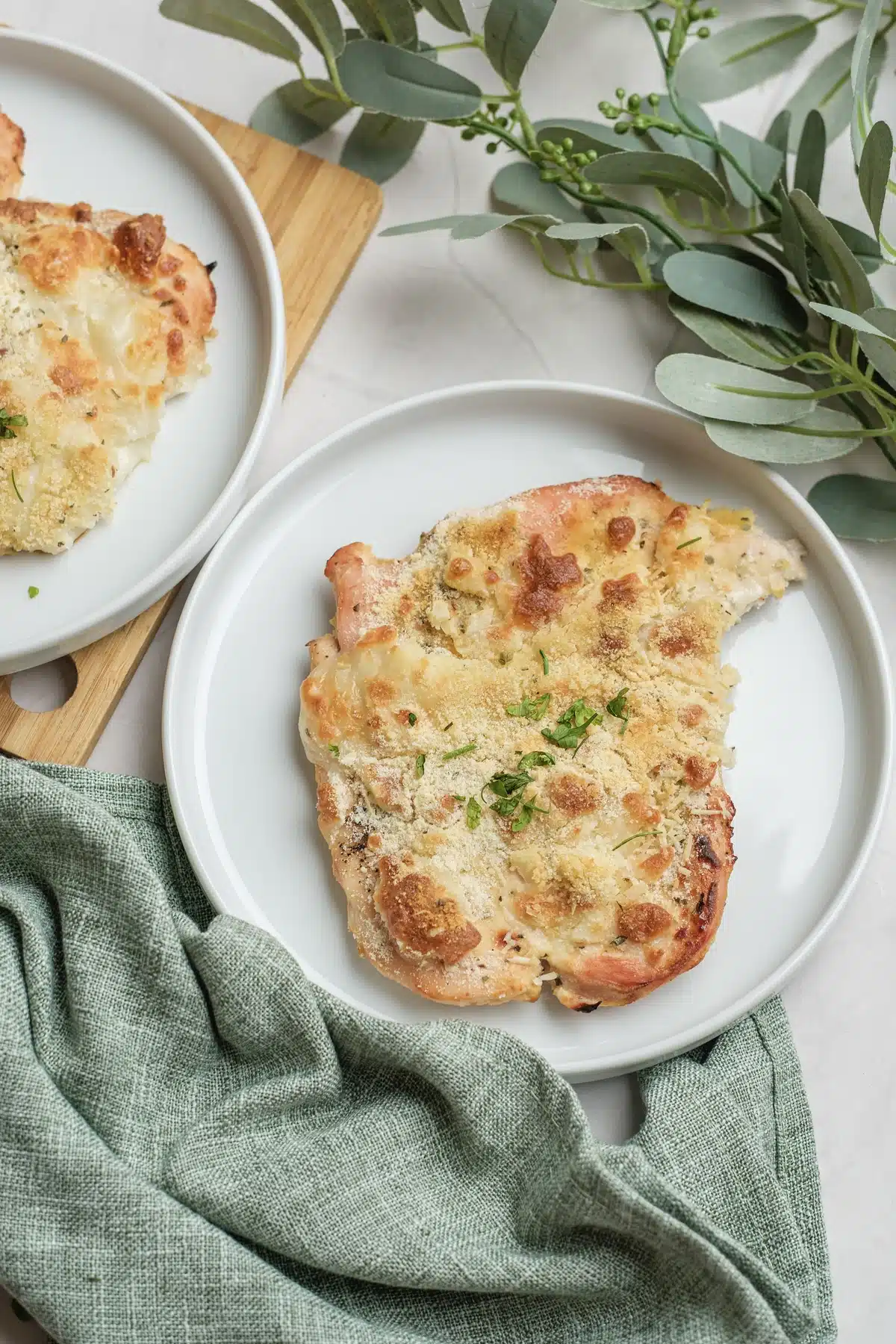 Baked Caesar chicken on two white plates with a golden cheese and breadcrumb top, parsley and a sprig of eucalyptus