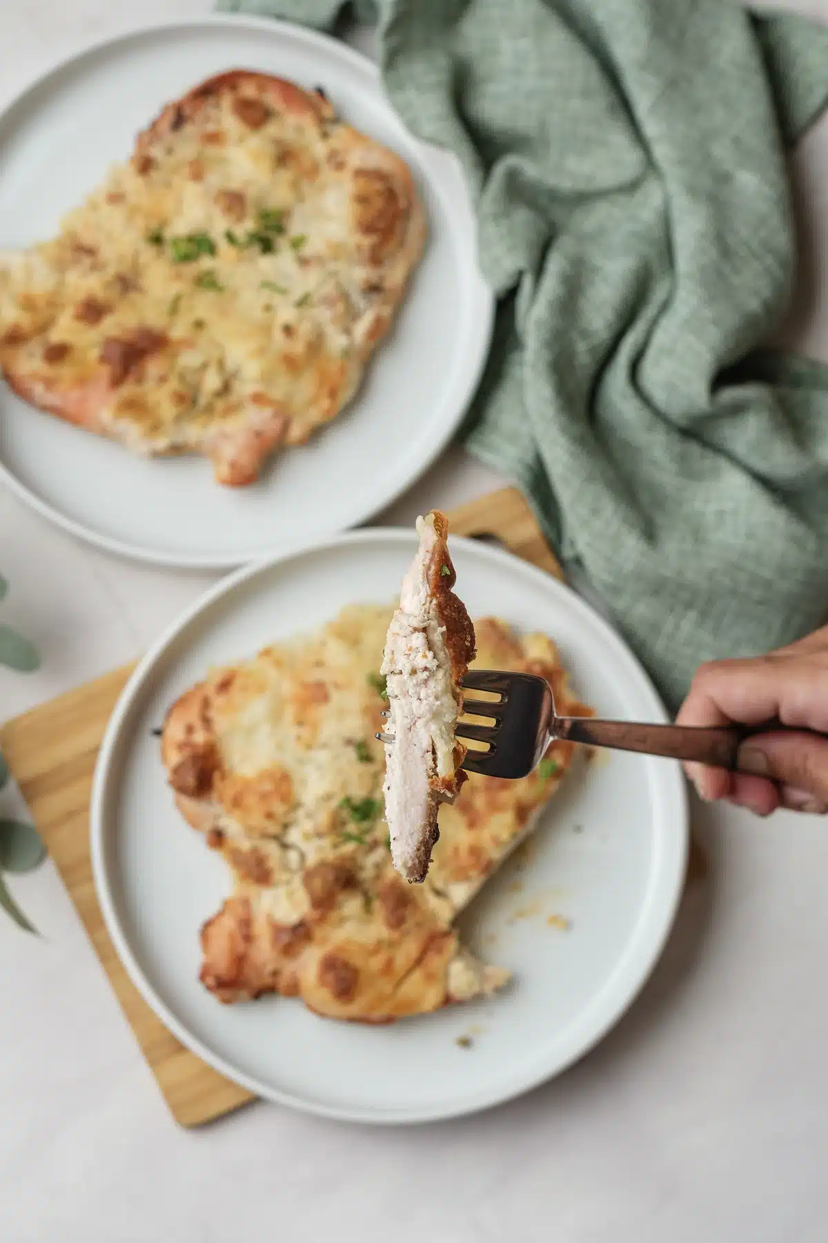 Caesar chicken on a white plate with a fork lifting a cut piece, a second plate and a green napkin behind