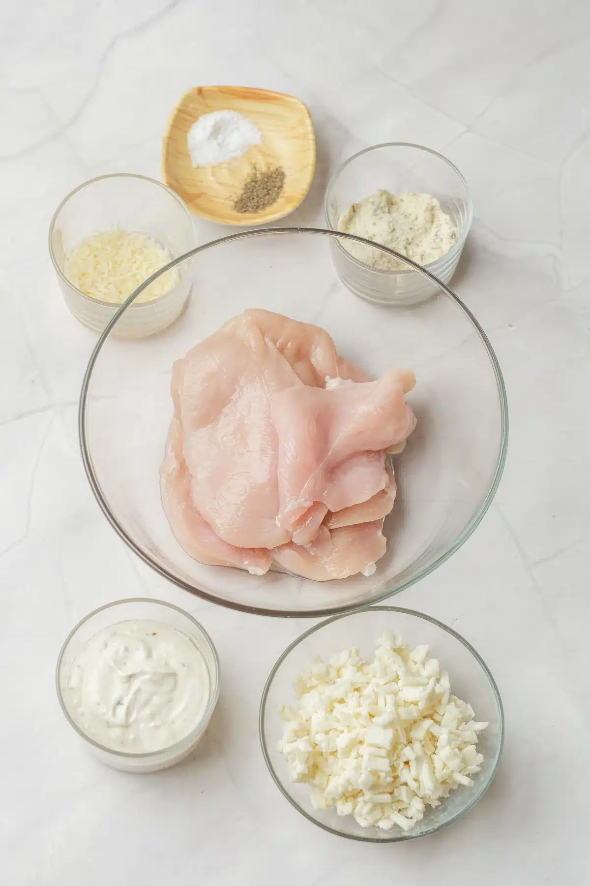 Raw breasts in a glass bowl surrounded by dishes of creamy dressing, shredded mozzarella, grated Parmesan, breadcrumbs, salt and pepper