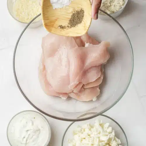 Salt and pepper sprinkled from a small wooden dish over raw breasts in a glass bowl