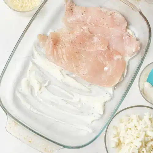 A raw breast laid on the dressing in the glass baking dish, with bowls of cheese beside it