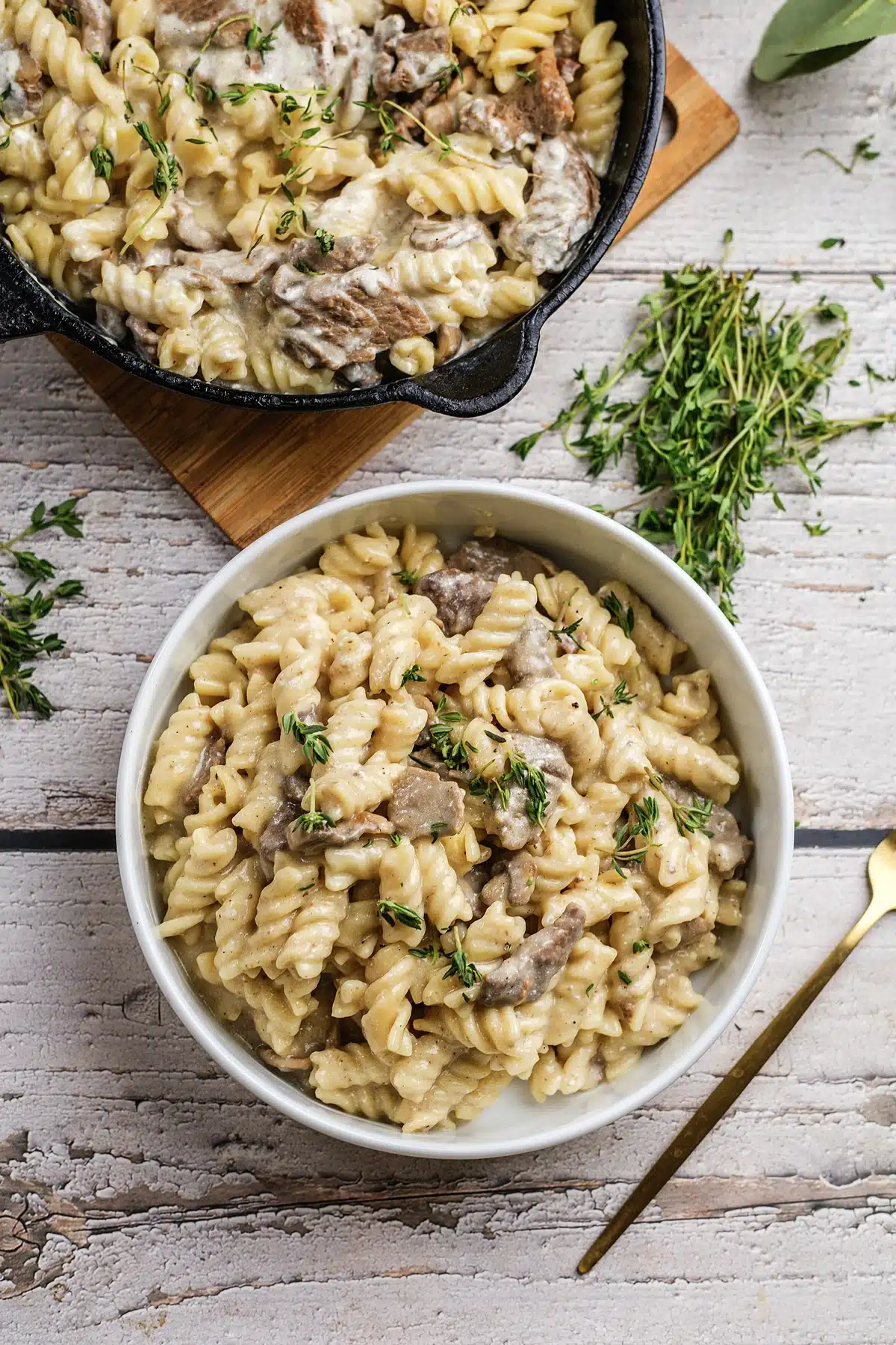 Beef stroganoff in a white bowl with the skillet and fresh thyme alongside