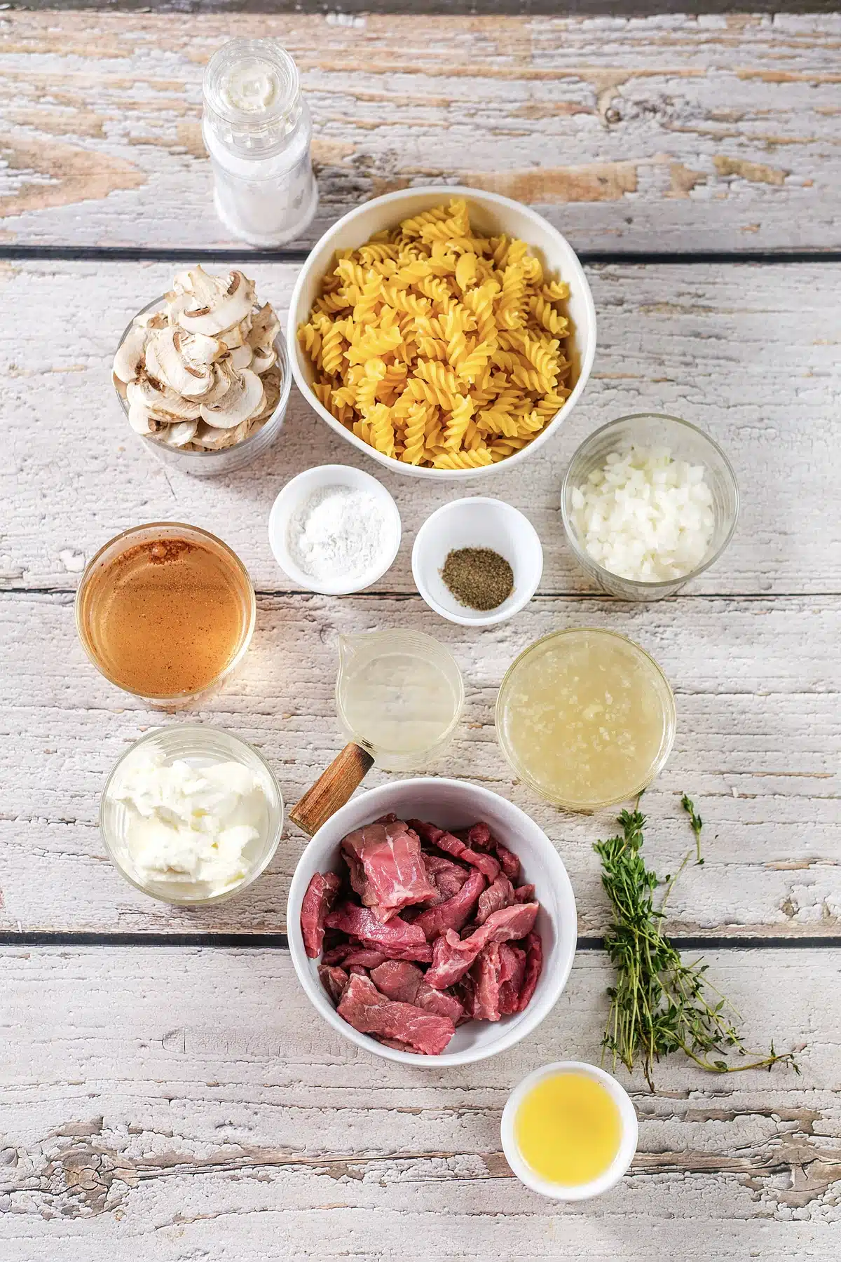 Sliced steak, mushrooms, dry pasta, sour cream, stock and thyme laid out on white wood