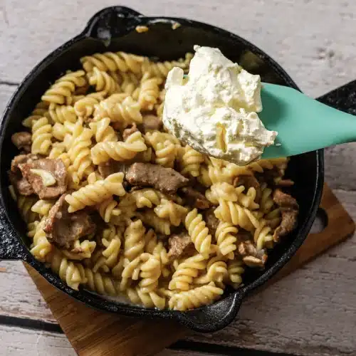 A spatula lowering a spoonful of sour cream into the cooked pasta