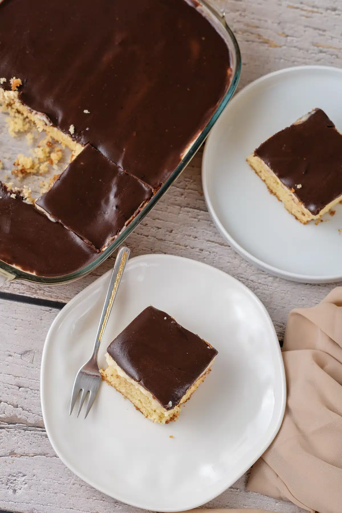 Boston cream poke cake in its glass dish from above with squares cut and one lifted out, beside a white plate holding a serving