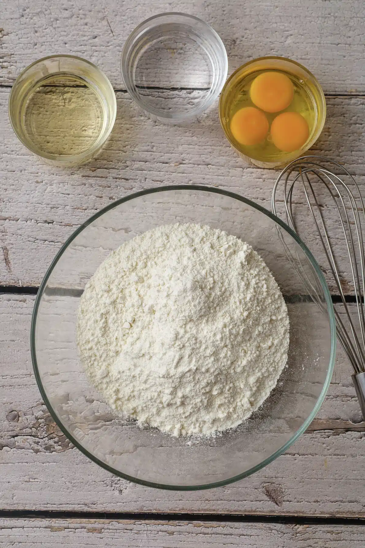 A bowl of yellow mix with three eggs, a cup of oil, a cup of water and a whisk laid out on a white board