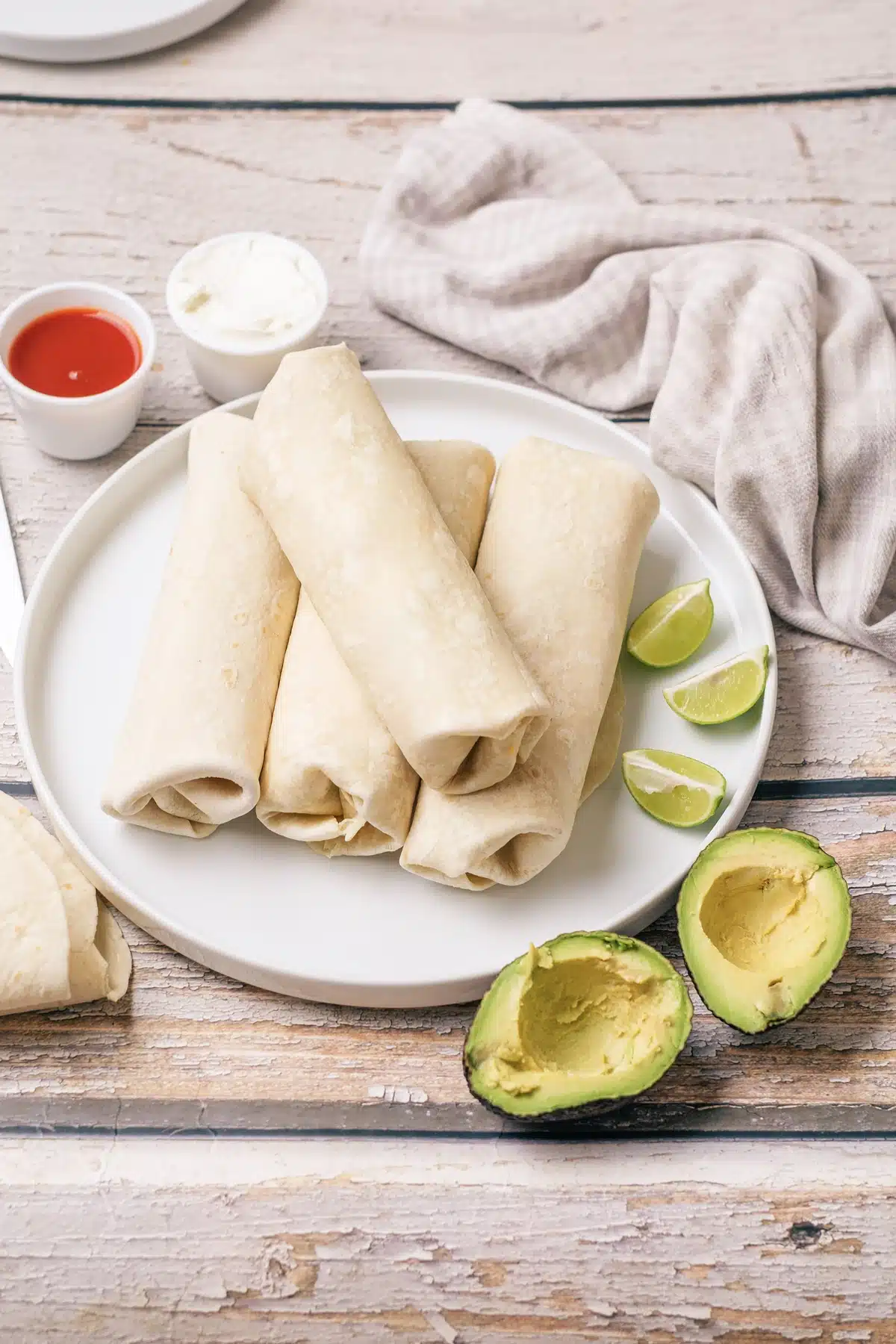 Six breakfast burritos rolled and stacked on a white plate with lime wedges, avocado halves, hot sauce and sour cream