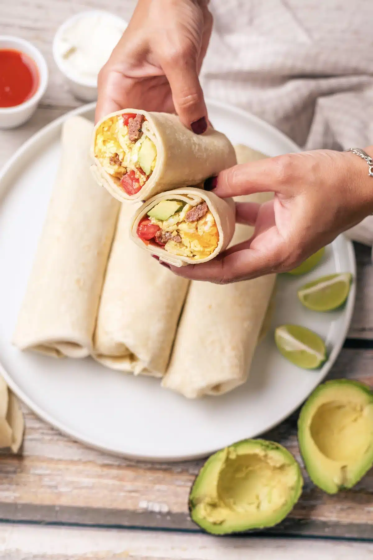 Hands holding a cut breakfast burrito above the stacked plate, with sour cream and hot sauce behind