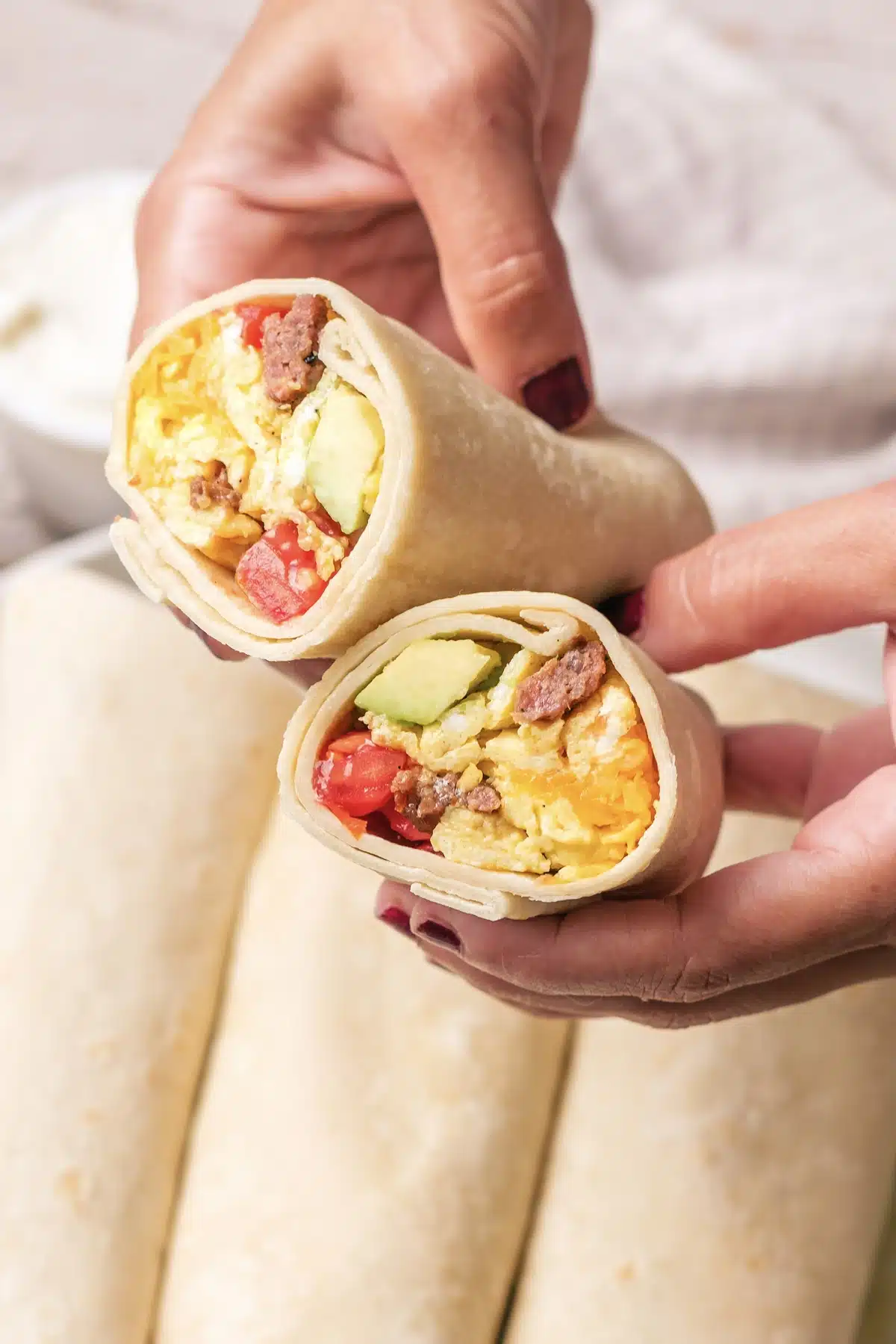 Two hands holding the cut halves of a wrap to show the scrambled egg, sausage, tomato and avocado filling