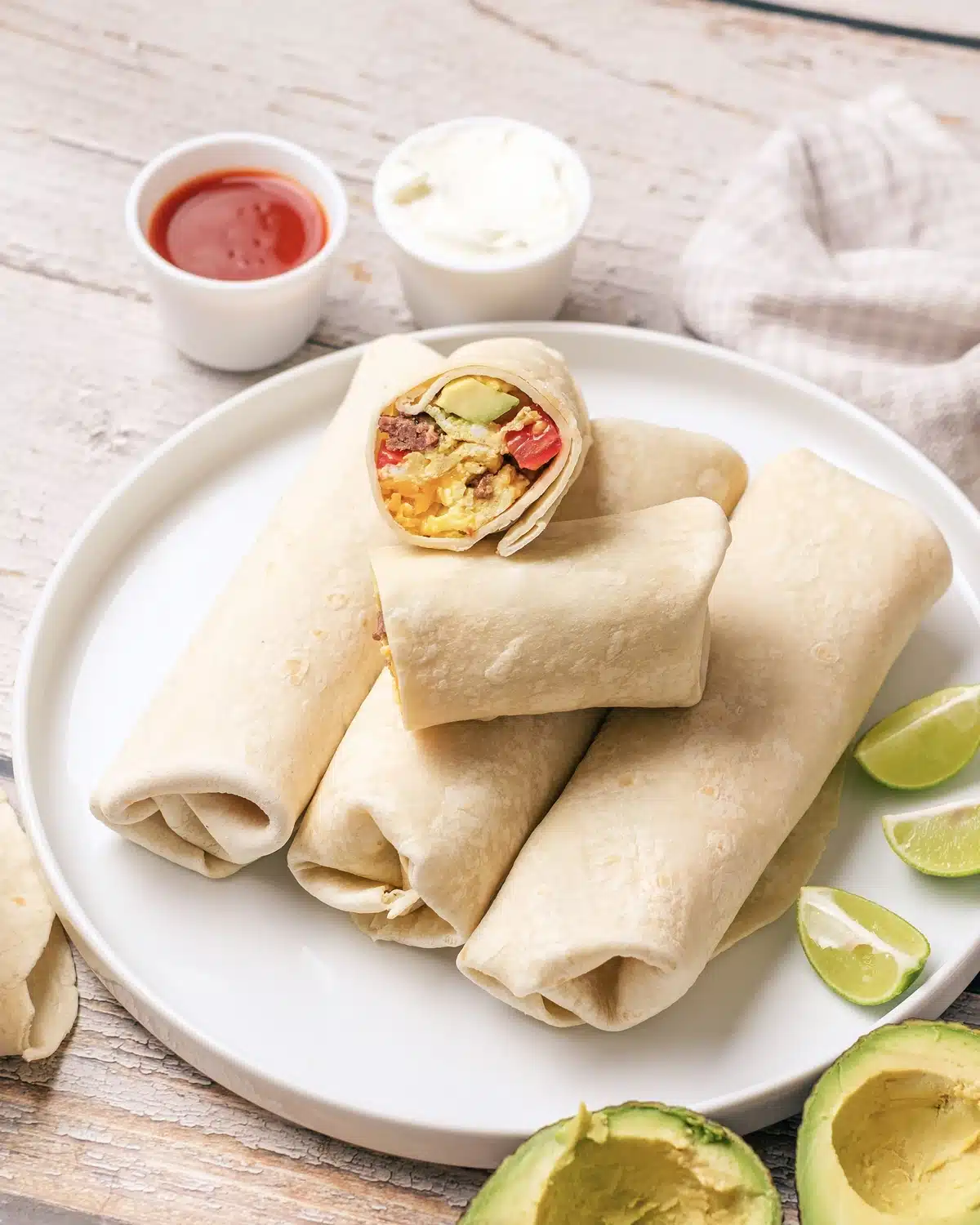 Breakfast burritos on a plate with one cut in half on top to show the egg, sausage, tomato and avocado inside