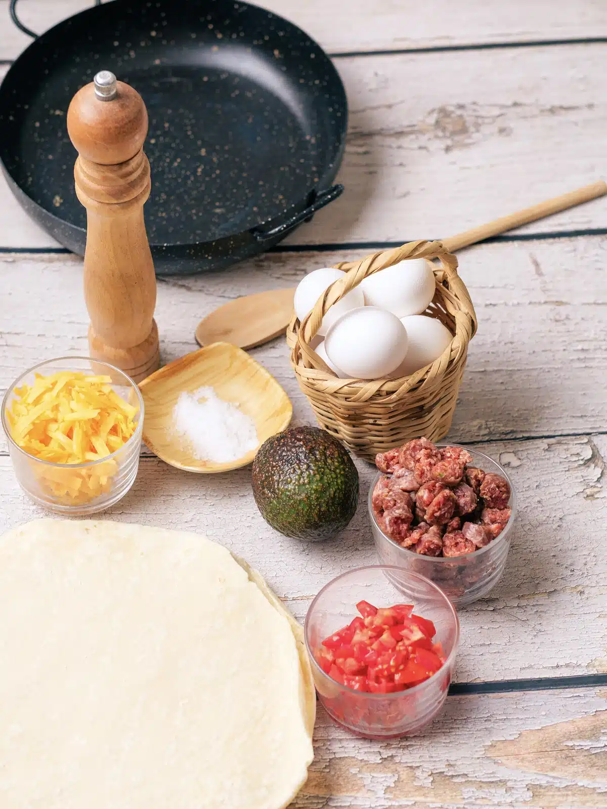 A skillet, eggs in a basket, shredded cheddar, cooked sausage crumbles, diced tomato, an avocado, salt and flour tortillas on a wooden table