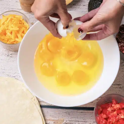Eggs cracked into a white bowl beside a flour tortilla and a bowl of diced tomato