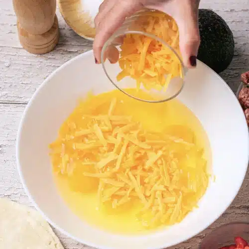 Shredded cheddar tipped from a glass into the bowl of eggs