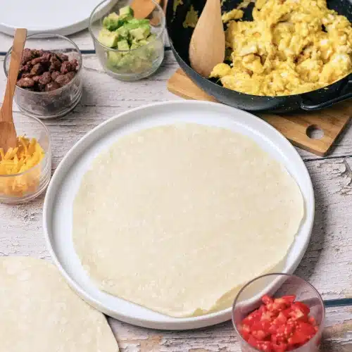 Soft scrambled eggs in a skillet with a wooden spoon, a tortilla on a plate and bowls of sausage and avocado beside