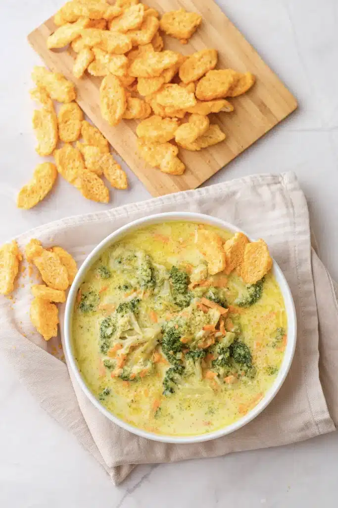 Broccoli cheddar soup in a white bowl seen from directly above, with a board of cheese crackers at the top of the frame