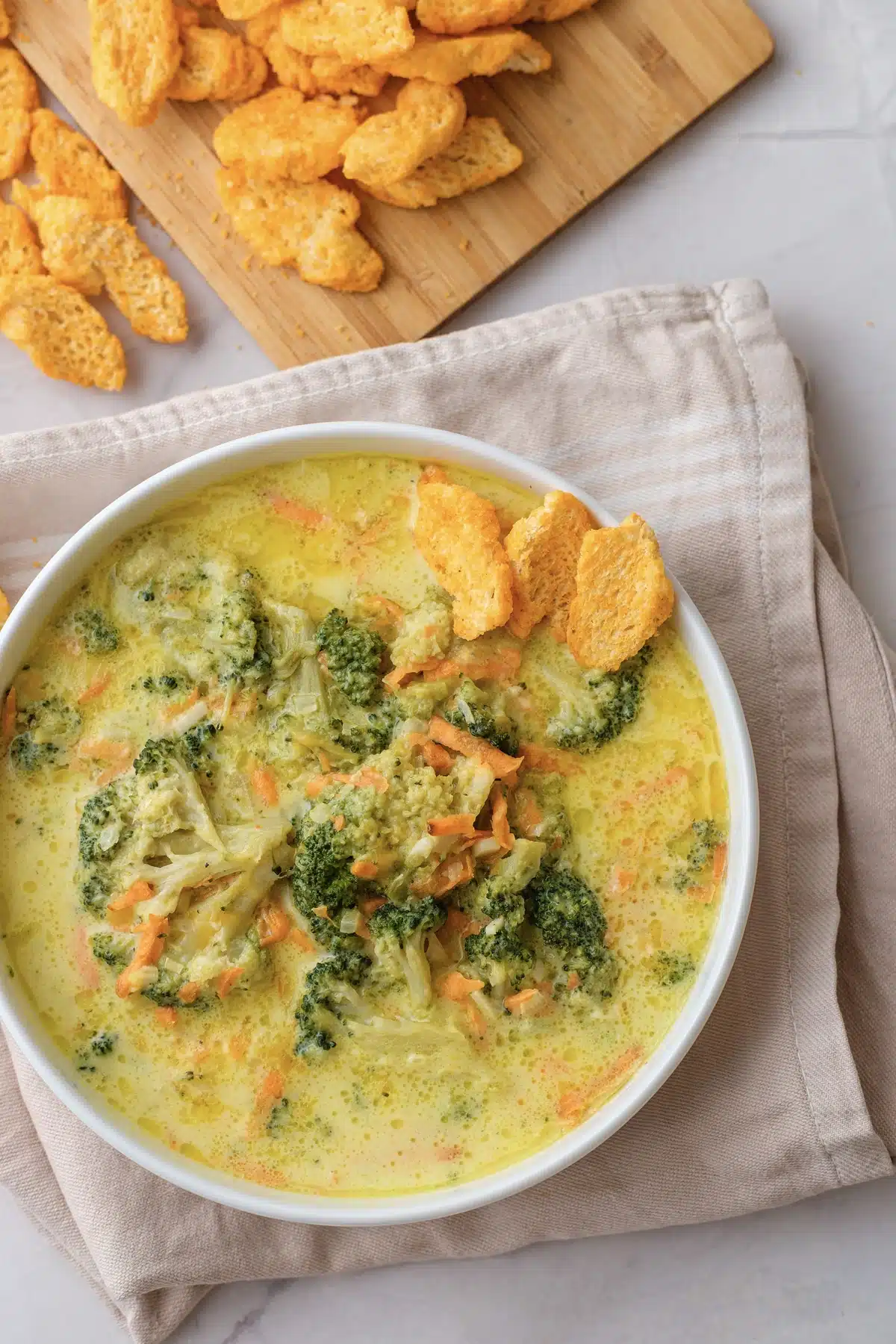 Broccoli cheddar soup in a white bowl with whole florets, shredded carrot and a few cheese crackers on top