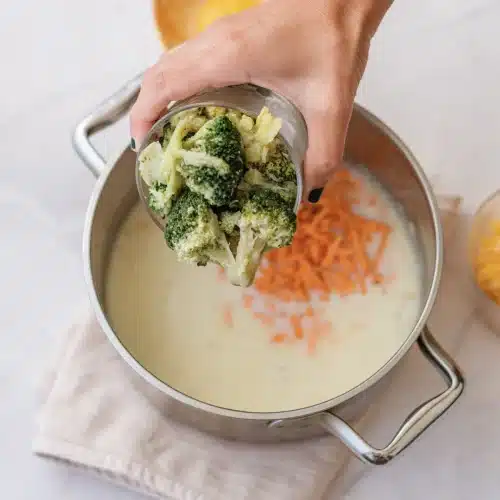 A hand adding broccoli florets and shredded carrot to the pot