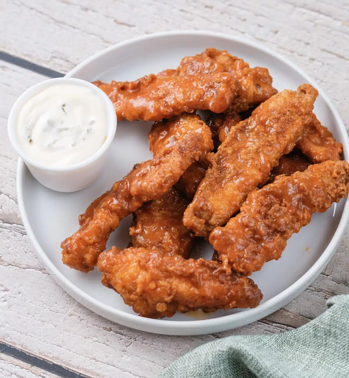 Oven-baked buffalo chicken tenders piled on a white plate with a cup of ranch dip and a green napkin
