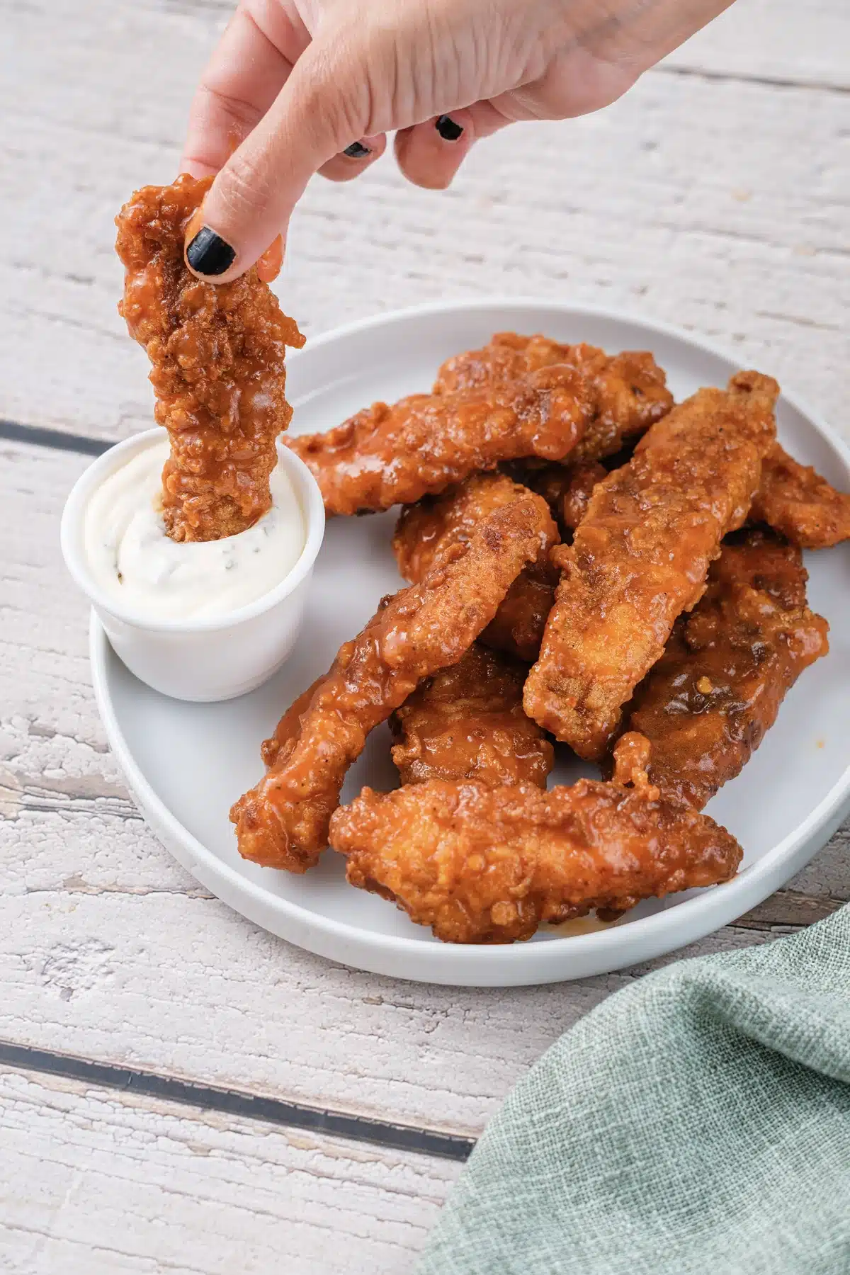 A hand dipping a sauced tender into a cup of ranch above a plate of the rest