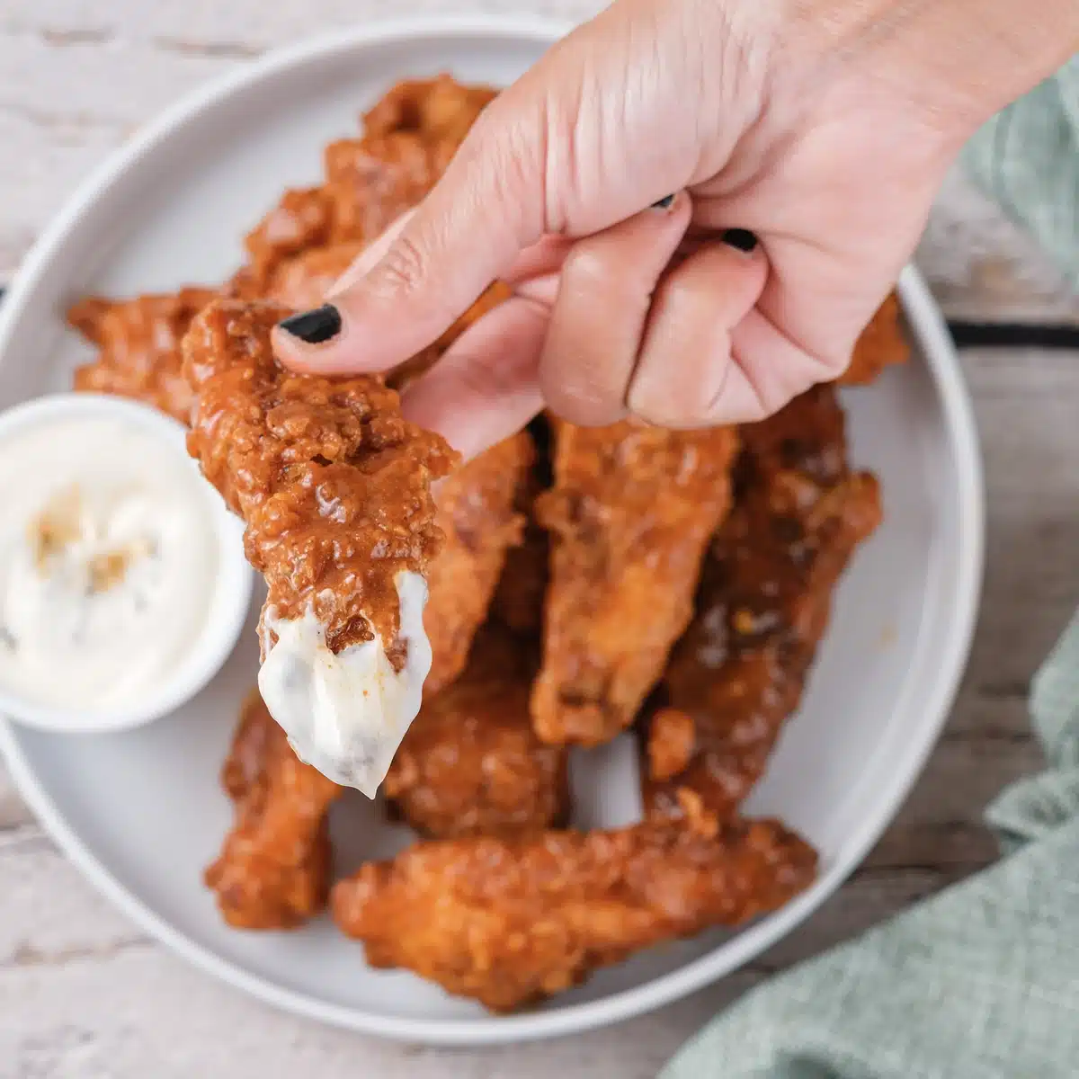 A hand holding a tender coated in orange sauce over a cup of ranch with a plate below