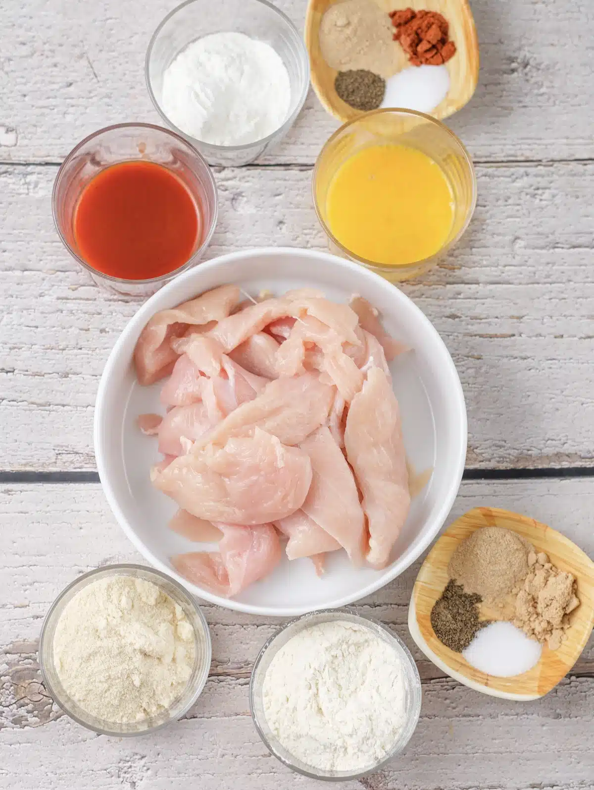 Raw chicken strips in a bowl with hot sauce, beaten egg, flour, cornstarch, fine breadcrumbs and a dish of spices