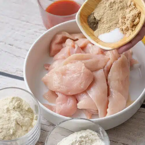 Salt and spices tipped from a small dish over raw chicken strips in a white bowl